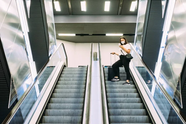 Yüz maskeli genç turist akıllı telefonunu kullanırken metro yürüyen merdivenini kullanıyor.
