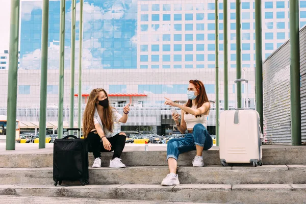 Yüz maskesi takan iki genç turist covid-19 'dan güvenli bir mesafede oturuyorlar ve akıllı telefonlarıyla otele giden yolu arıyorlar.