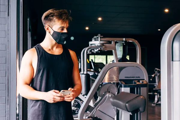Yüz maskesi takan genç adam spor salonunda akıllı telefonuna bakıyor.
