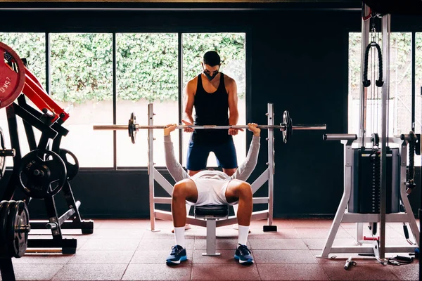 Yüz maskesi takan kişisel antrenör, genç erkeklerin spor salonunda egzersiz yapmalarına yardımcı oluyor.