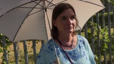 Güneşli bir günde elinde güneş şemsiyesi tutan orta yaşlı bir kadının samimi portresi. Gülümsüyor, bakışlarını kaçırıyor, yaz sıcağının tadını çıkarıyor.