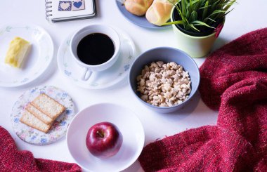 Beyaz yemek masasındaki kahvaltı menüsünün en üst görüntüsü. Kahve, süt, mısır gevreği, elma, kraker ve çörekler yukarıdan beyaz masa manzaralı..
