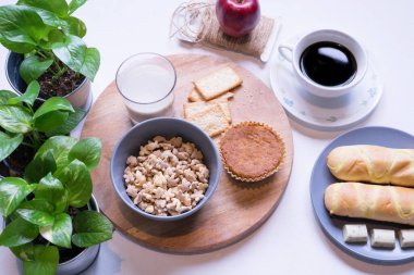 Beyaz yemek masasındaki kahvaltı menüsünün en üst görüntüsü. Kahve, süt, mısır gevreği, elma, kraker ve çörekler yukarıdan beyaz masa manzaralı..
