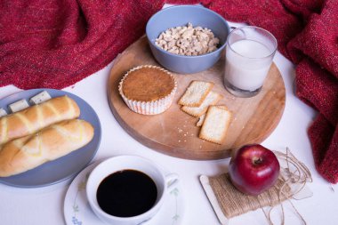 Beyaz yemek masasındaki kahvaltı menüsünün en üst görüntüsü. Kahve, süt, mısır gevreği, elma, kraker ve çörekler yukarıdan beyaz masa manzaralı..