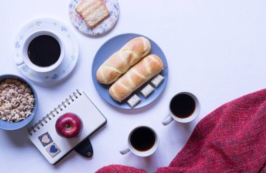 Beyaz yemek masasındaki kahvaltı menüsünün en üst görüntüsü. Kahve, süt, mısır gevreği, elma, kraker ve çörekler yukarıdan beyaz masa manzaralı..