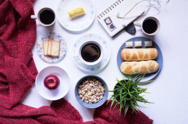 Beyaz yemek masasındaki kahvaltı menüsünün en üst görüntüsü. Kahve, süt, mısır gevreği, elma, kraker ve çörekler yukarıdan beyaz masa manzaralı..