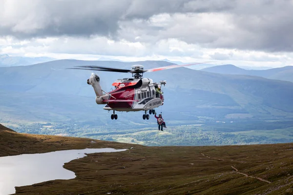 Helikopter Ben Nevis 'in inişi sırasında yere düşen bir turisti uçurmak için geldi. Resimde helikopterin altındaki ipte asılı iki kişi görüyoruz. Arka planda vadi ve diğer dağlar var..