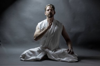 Meditasyon pozu veren bir adam, bir kolu havada, gri arka planda.