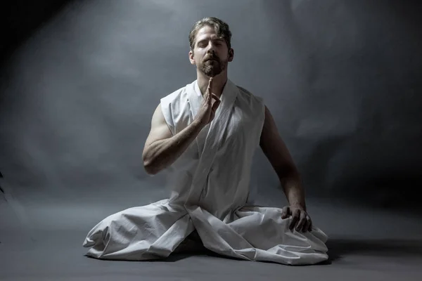 Meditasyon pozu veren bir adam, bir kolu havada, gri arka planda.