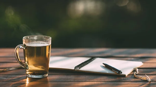 Transparent cup with hot tea with steam and opened paper notebook for planning with pen on textured wooden table made of aged boards against blurred background of evening garden. Concept of relaxation, creativity, tranquility.