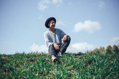 şapka giyen genç adam modeli çimenlerin üzerinde oturan ve poz o moda geri kalan geçiriyor