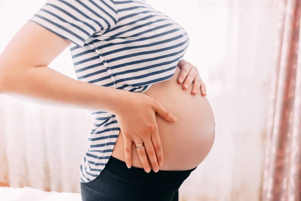 シャツで妊娠中の女性は ウィンドウに対して自宅で白い背景の上の腹に手を保持します 出産準備 期待のコンセプトです クローズ アップ コピー スペース 屋内で 妊娠の美しい柔らかい雰囲気写真 — ストック写真