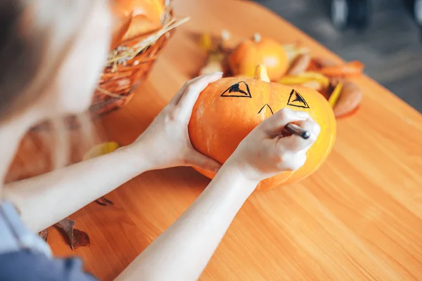 Woman hands drawing scary face on pumpkin with a black marker pen on home background for Halloween. Holiday decoration concept.