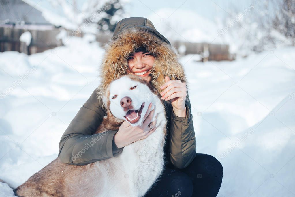 A young woman play with the dog red husky with blue eyes. Snowy morning in the garden. A girl dressed in warm clothes