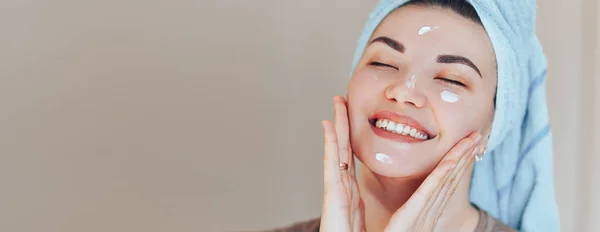 Happy smiling pretty beautiful woman girl with towel on head smiling touch healthy clean soft moisturized hydrated skin care after applying cream on young face — Stock Photo, Image