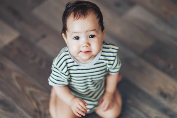 Beautiful smiling baby girl with big dark eyes dark hair sitting at floor and looking up at camera — Stock Photo, Image