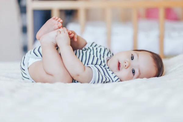 Glückliches Baby auf weißem Laken liegend und ihre Beine haltend. verspieltes Baby im Bett liegend — Stockfoto