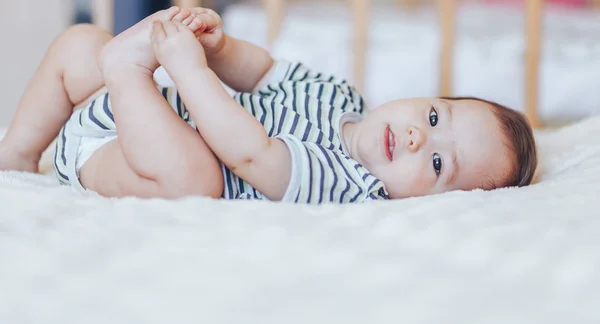 Happy baby lying on white sheet and holding her legs. Playful baby lying down in bed — Stock Photo, Image