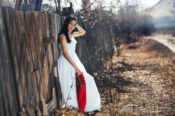 Portrait d'une belle fille aux cheveux noirs dans une robe vintage blanche debout près d'une clôture en bois.Modèle jeune femme posant dans un style national russe. châle rouge — Photo