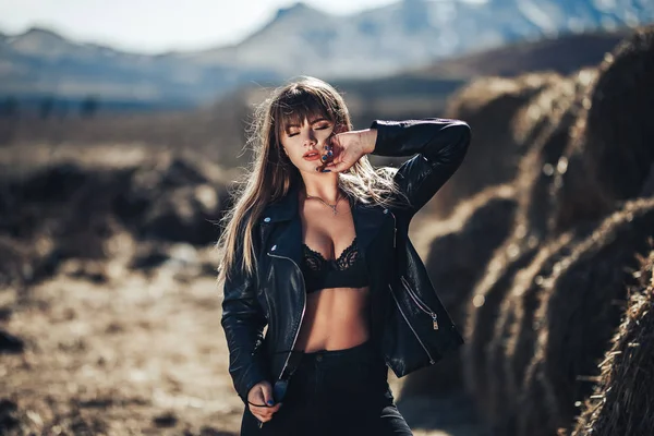 Сексуальна красива молода жінка в чорному бюстгальтері, шкіряній куртці та чорних шкіряних штанях позує біля сонячного світла сіна — стокове фото