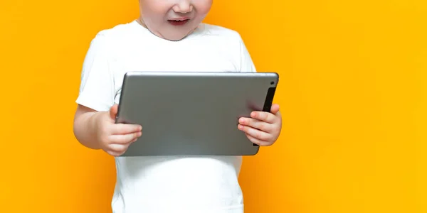 Surpreso loira de três anos de idade menino com a boca aberta surpreso, segurando em suas mãos um tablet pc e olhando para a câmera no fundo amarelo — Fotografia de Stock