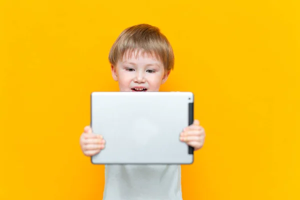 Sorprendido rubia de tres años de edad, chico con la boca abierta sorprendido, sosteniendo en sus manos una tableta de PC y mirando a la cámara sobre fondo amarillo — Foto de Stock