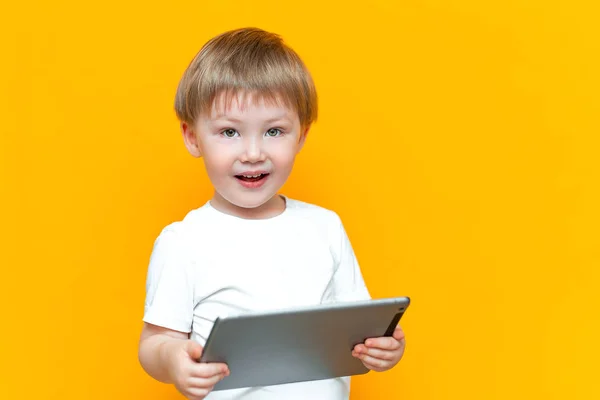 Sorprendido rubia de tres años de edad, chico con la boca abierta sorprendido, sosteniendo en sus manos una tableta de PC y mirando a la cámara sobre fondo amarillo — Foto de Stock