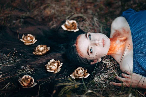 Porträt einer schönen, braunhaarigen Frau in einem blauen Kleid im Gras liegend, mit goldenen Rosen auf ihren langen Haaren, im Märchenwald, goldener Farbschimmer am Hals Fantasiekonzept — Stockfoto