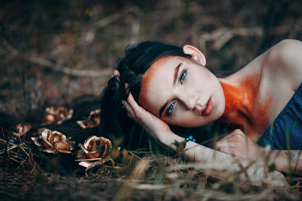 Portrait of beautiful brown-haired woman in a blue dress lying on grass, with golden roses on her long hair, in the fairy forest, golden paint shimmer on her neck fantasy concept.