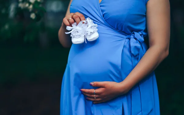 Embarazada mujer sosteniendo blanco babys bootee usando en vestido azul sobre fondo del parque natural al atardecer . — Foto de Stock