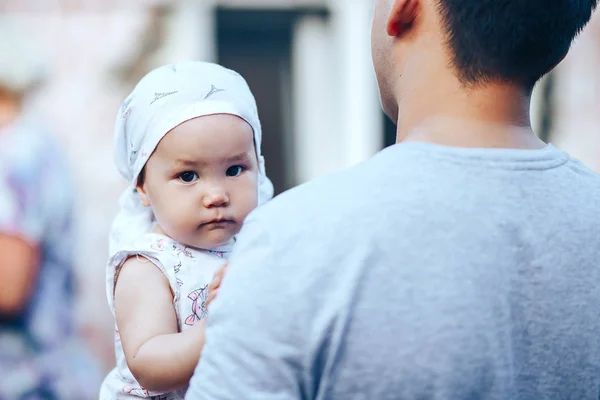 Kleine donkerharige baby meisje op vaders handen buiten, kijken naar camera — Stockfoto