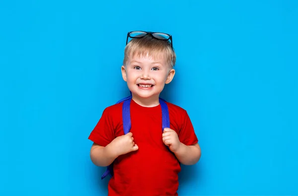 Menino sorridente feliz em camiseta vermelha com óculos na cabeça vai para a escola pela primeira vez. Criança com saco escolar. Miúdo no fundo azul. De volta à escola — Fotografia de Stock