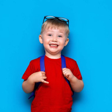 Kırmızı tişörtlü, kafasında gözlüklü mutlu gülümseyen çocuk ilk kez okula gidiyor. Okul çantası olan çocuk. Mavi arka planda çocuk. Okula geri dön