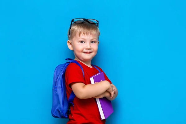 Mały szczęśliwy uśmiechnięty chłopak z okularami na głowie, książka w rękach, Torba szkolna na ramionach. Powrót do szkoły. gotowy do szkoły — Zdjęcie stockowe