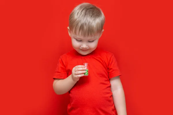Μικρό αγόρι ξανθά μαλλιά πρόκειται να φάει πολλά δισκία χάπια στα χέρια του, επικίνδυνο χωρίς ενήλικες ανθρώπους, κόκκινα ρούχα και κόκκινο φόντο — Φωτογραφία Αρχείου