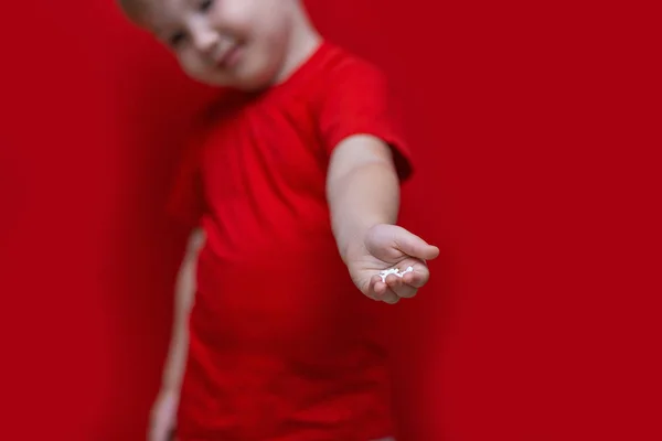 Μικρό λυπημένο παιδί κρατάει πολλά χάπια στα χέρια. κόκκινο μπλουζάκι, κόκκινο φόντο επικίνδυνη έννοια, δισκία και το μωρό — Φωτογραφία Αρχείου