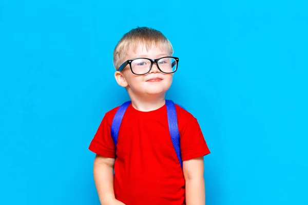 メガネをかけた赤いTシャツを着た幸せな笑顔の少年が初めて学校に行きます。スクールバッグを持つ子供。青い背景の子供。学校に戻る — ストック写真