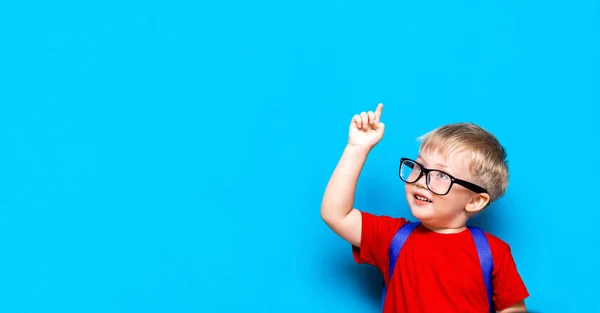 Back to school First grade junior lifestyle. Small boy in red t-shirt. Close up studio photo portrait of smiling boy in glasses with schoolbag and book show somethind with his finger — Stock Photo, Image
