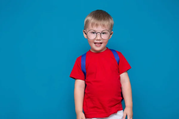 Το χαρούμενο χαμογελαστή αγόρι με το κόκκινο μπλουζάκι με τα στρογγυλά ποτήρια πάει στο σχολείο για πρώτη φορά. Παιδί με σχολική τσάντα. Το παιδί σε μπλε φόντο. Πίσω στο σχολείο — Φωτογραφία Αρχείου
