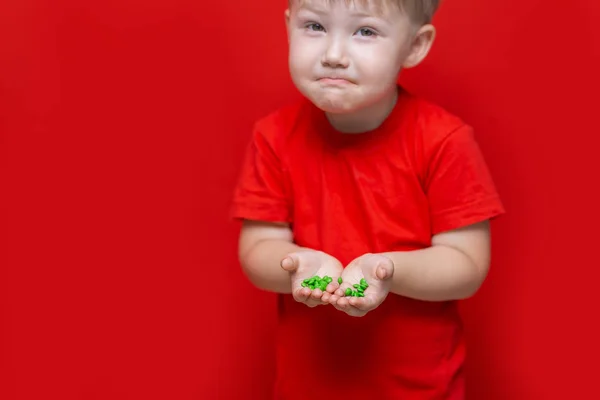Μικρό λυπημένο παιδί κρατάει πολλά χάπια στα χέρια. κόκκινο μπλουζάκι, κόκκινο φόντο επικίνδυνη έννοια, δισκία και το μωρό — Φωτογραφία Αρχείου