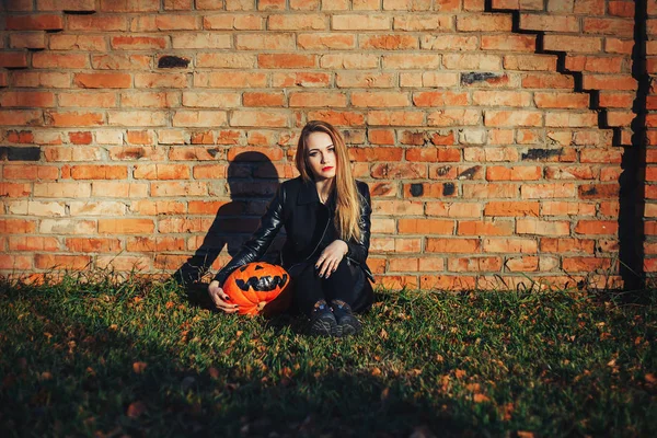 Mulher moderna de Halloween. Bruxa atraente segurando abóbora 'truque ou deleite'. Mulher vestindo jaqueta de couro e saia preta. Dia das Bruxas de Outubro. tijolos fundo — Fotografia de Stock