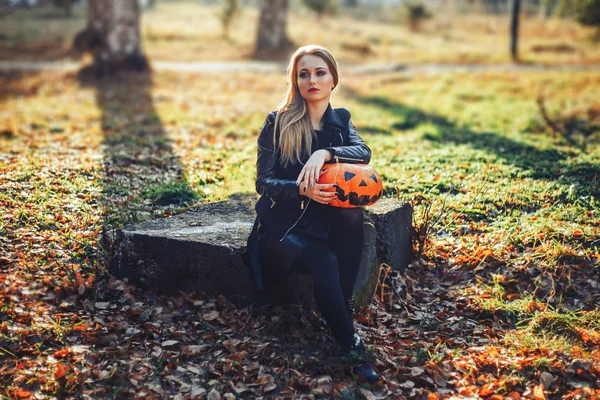 Belle jeune femme blonde avec un maquillage extravagant dans une veste en cuir noir avec les yeux grands ouverts et une bouche ouverte avec une citrouille dans les mains — Photo