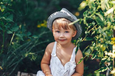 şapka ve rahat giysiler içinde sevimli çocuk domates ang kamera bakarak oturan, bahçede poz çocuk modeli