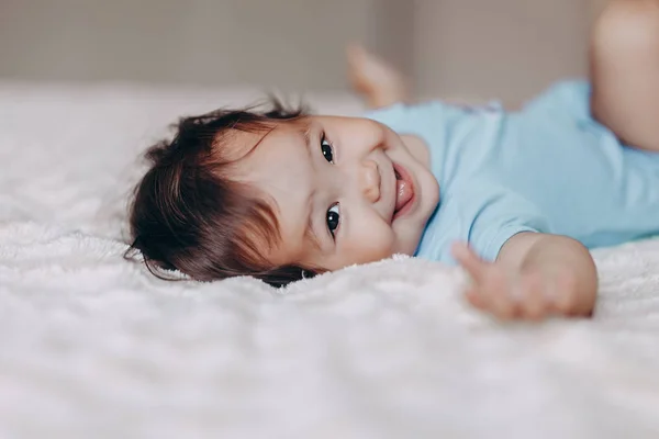 Cute laughing one year old girl lying on bed and looking at camera touch her feet — Stock Photo, Image