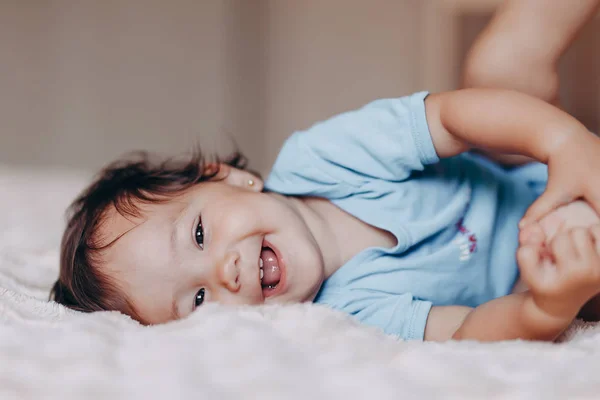 Cute laughing one year old girl lying on bed and looking at camera touch her feet — Stock Photo, Image