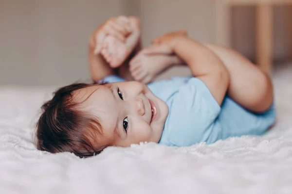 Süßes, lachendes einjähriges Mädchen, das auf dem Bett liegt und in die Kamera schaut, berührt ihre Füße — Stockfoto