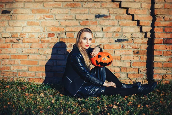 Mulher moderna de Halloween. Bruxa atraente segurando abóbora 'truque ou deleite'. Mulher vestindo jaqueta de couro e saia preta. Dia das Bruxas de Outubro. tijolos fundo — Fotografia de Stock