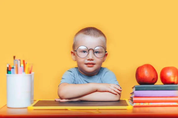 Lycklig blondin Smart pojke sitter vid ett skrivbord i runda glas och leende. Redo för skolan. Tillbaka till skolan. Äpple, pennor och böcker på skrivbordet — Stockfoto