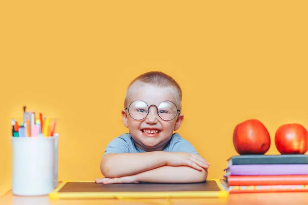 Lykkelig blond klog dreng sidder ved et skrivebord i runde briller og smiler. Klar til skole. Tilbage til skolen. Apple, kuglepenne og bøger på skrivebordet - Stock-foto