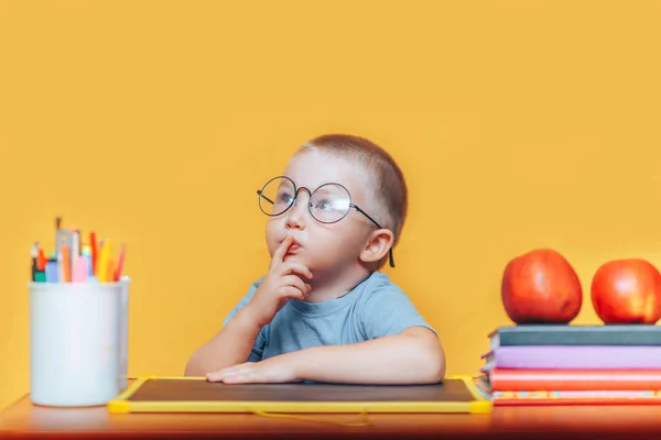 Хлопчик в круглих окулярах у сорочці і сидить за столом і думає, роблячи домашнє завдання, на жовтому тлі. назад до школи — стокове фото